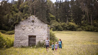 Familie wandert auf einer Wiese vor einer alten Steinhütte im Wald