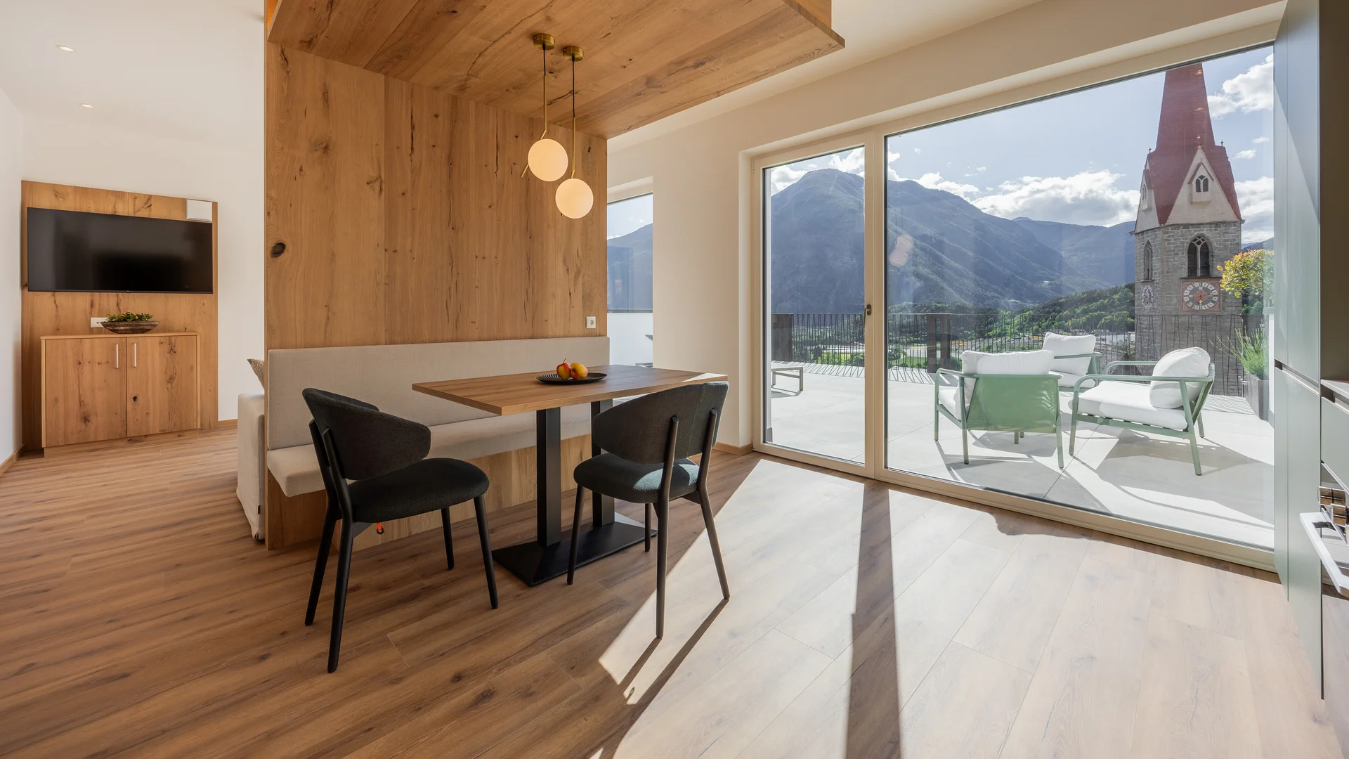 Living area with wooden furniture and terrace overlooking mountains and church tower