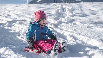 Kind sitzt auf einem Schlitten im Schnee vor einer verschneiten Bergkulisse