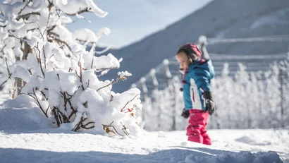 Kind in Winterkleidung neben schneebedeckten Sträuchern in einer verschneiten Berglandschaft