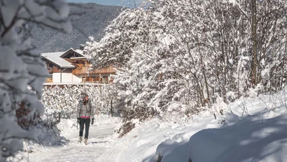 Frau spaziert auf einem verschneiten Weg zwischen schneebedeckten Bäumen und einem Berghaus