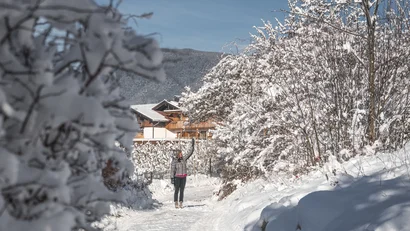 Person auf einem verschneiten Weg zwischen schneebedeckten Bäumen und einem Chalet