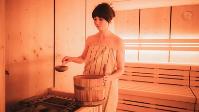 Frau mit Handtuch in Sauna gießt Wasser auf heiße Steine