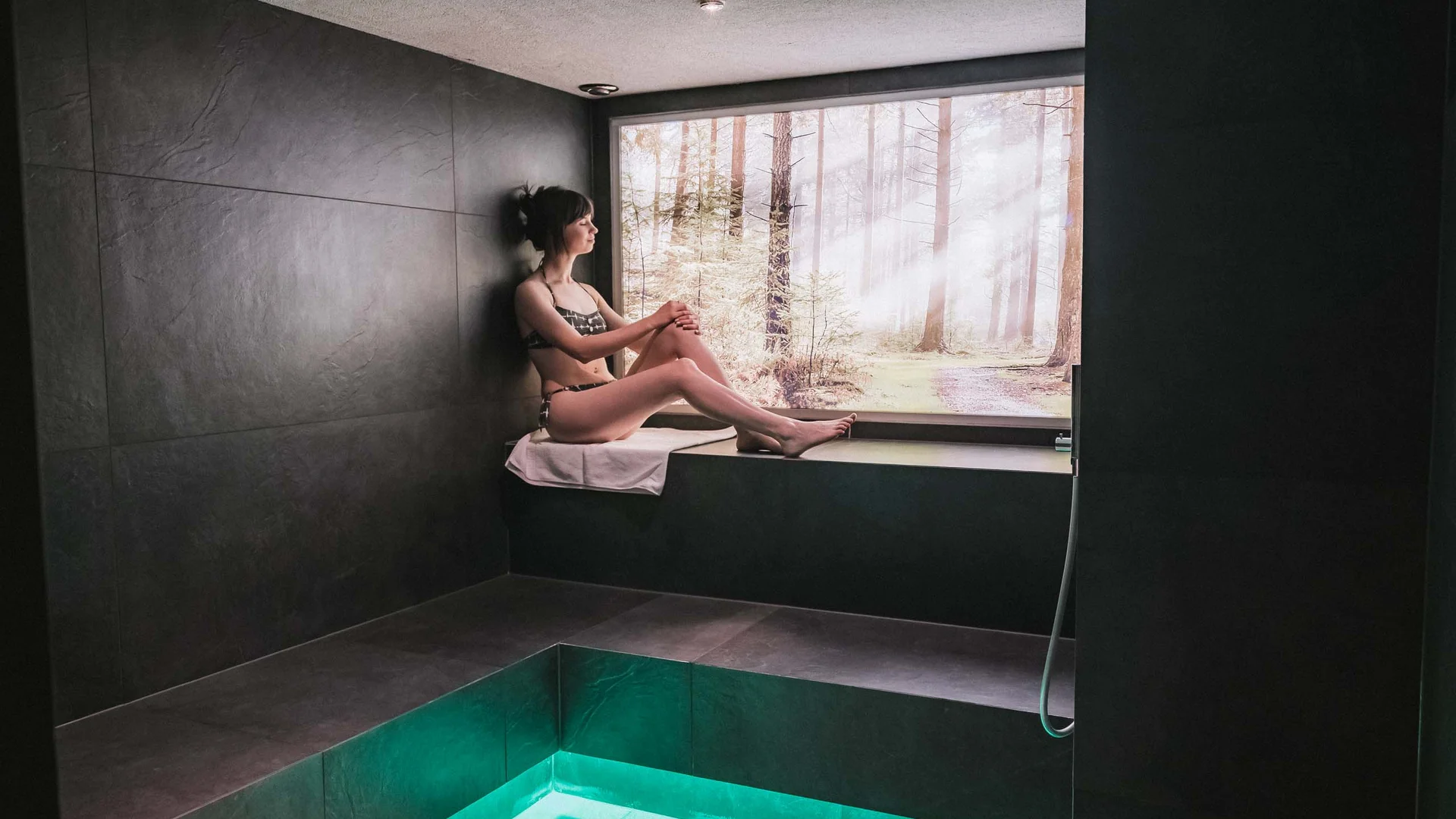 Frau in Bikini sitzt in dunklem Spa-Raum vor Fenster mit Waldblick