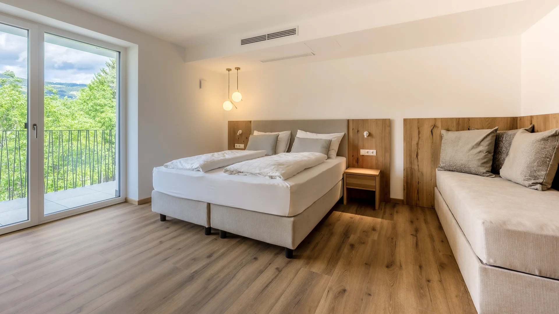 Helles Hotelzimmer mit Doppelbett, Sofa und Balkon mit Blick auf grüne Bäume