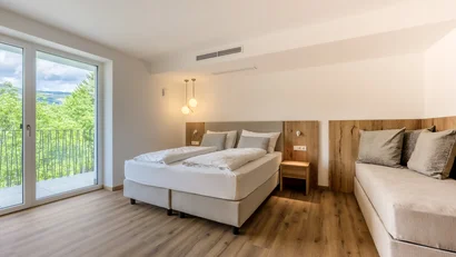 Bright hotel room with double bed, sofa, and balcony overlooking green trees